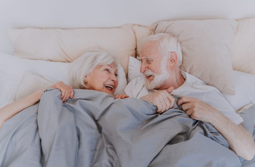 A senior couple nestled in bed, sharing warm smiles as they gaze at each other, with the blankets gently tucked up to their chins.