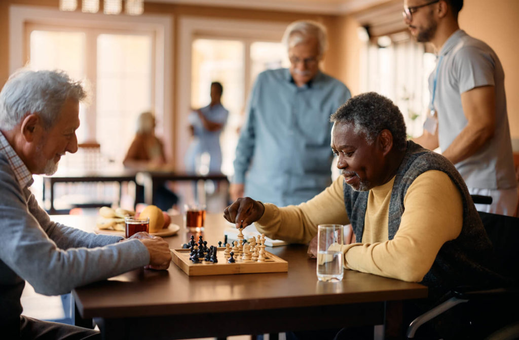 A pair of older adults in senior living playing chess in a common area and smiling.