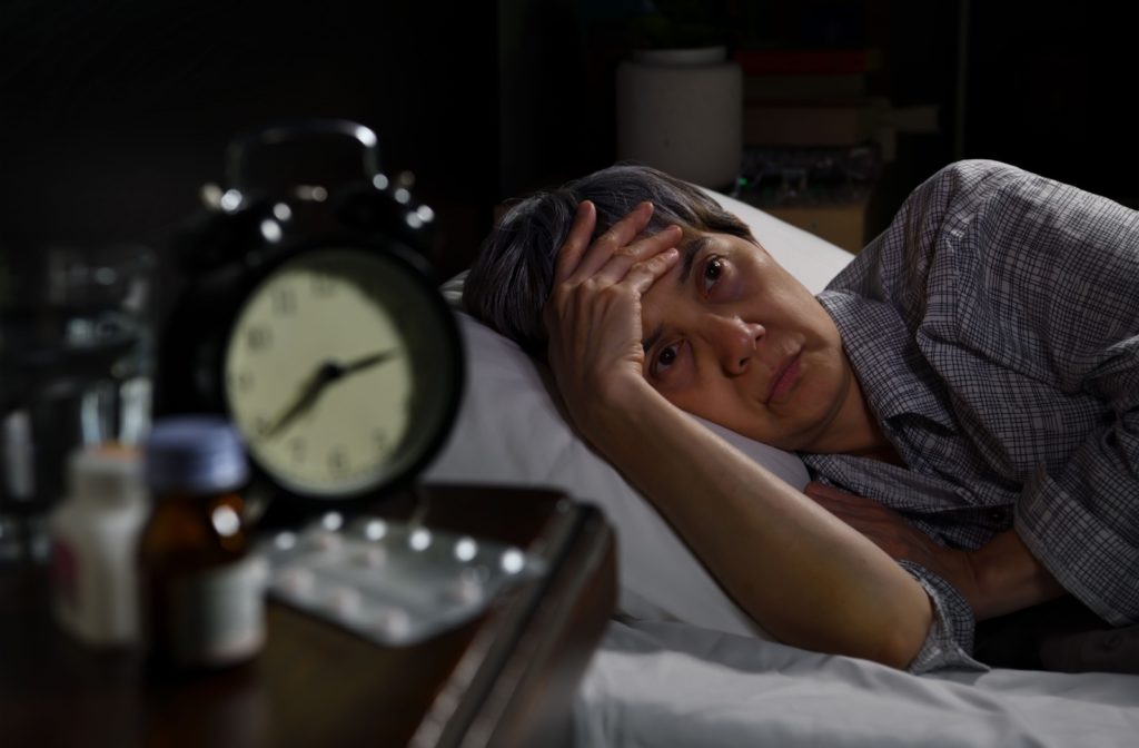 A senior woman lies in bed with a weary expression, her hand resting on her forehead. On the bedside table in the foreground, a clock and several medication bottles, including sleeping pills, hint at her restless nights.