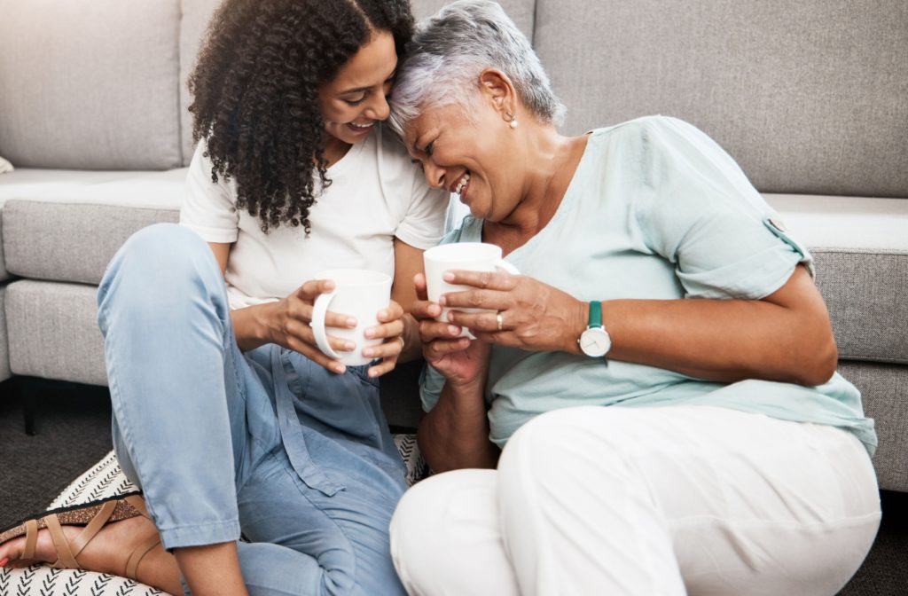 A daughter and senior mother sharing a cup of coffee and laughing together in a living room.