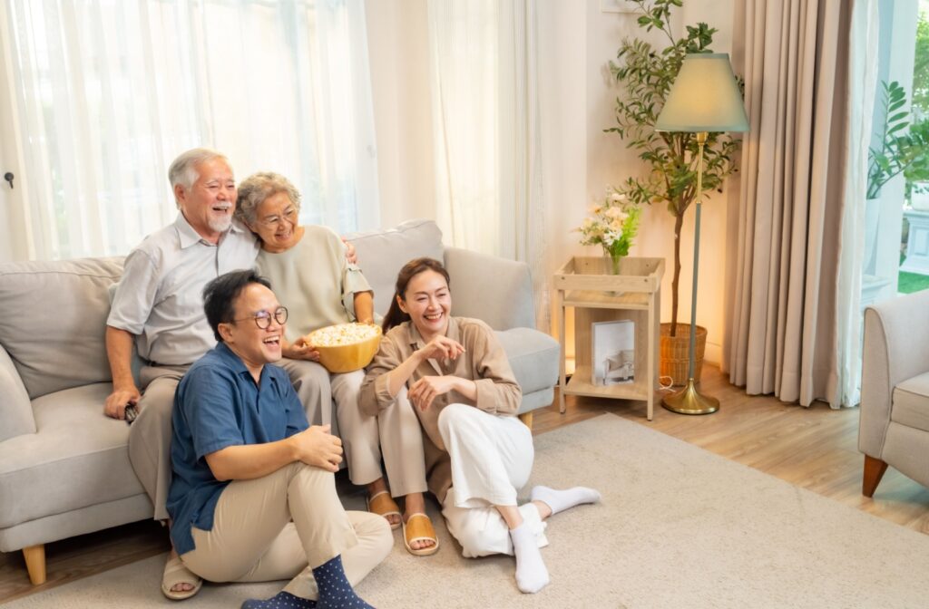 Two adults sit at their senior parent’s feet while the family giggles at a light-hearted part of a movie about dementia