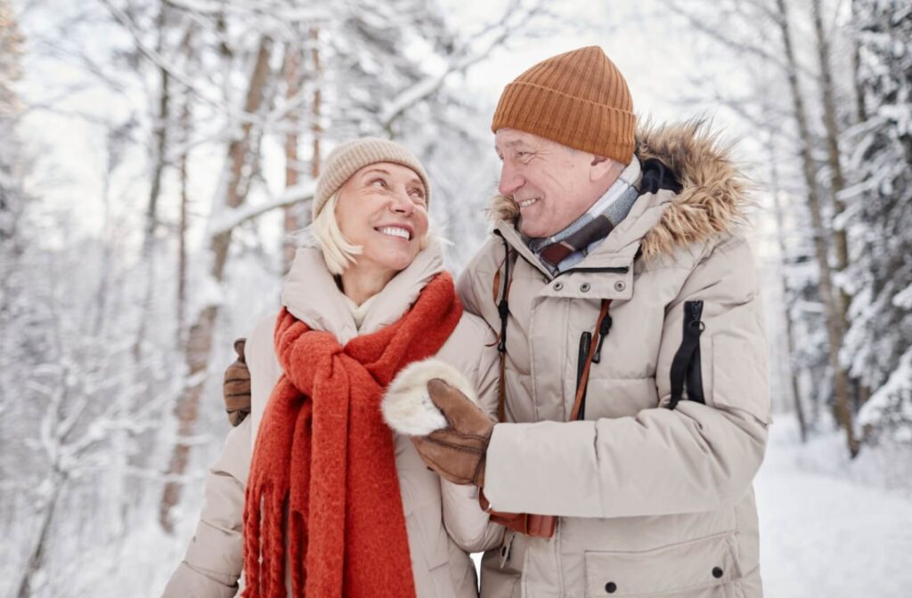 A senior couple in warm winter clothes hold each other and smile while enjoying a safe walk in a snowy winter forest