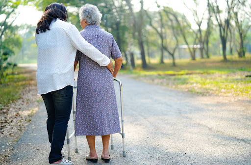 A younger woman supports a senior woman walking with a walker on a path surrounded by grass and trees.