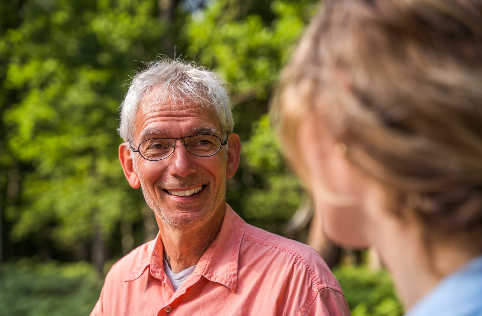 A senior man wearing glasses and smiling as he looks at a woman who is out of focus, facing away from the camera on the right side of the frame.