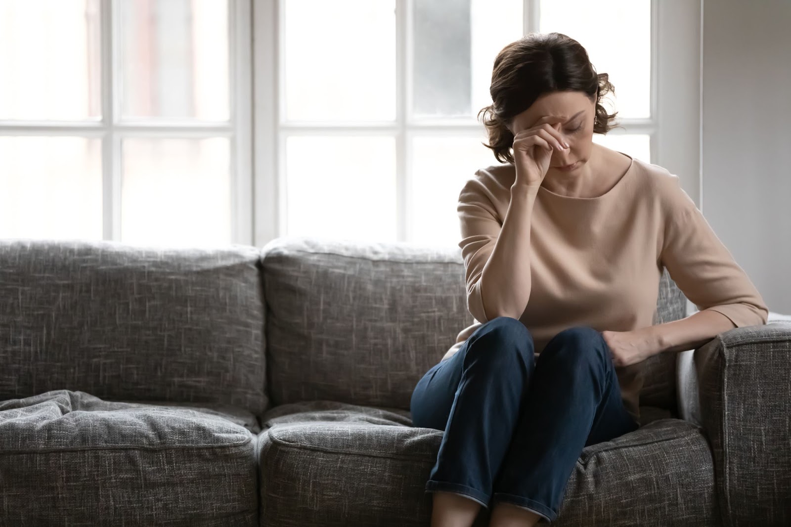 A tired looking woman sitting on the couch struggling with caregiver burnout from caretaking her senior parent.