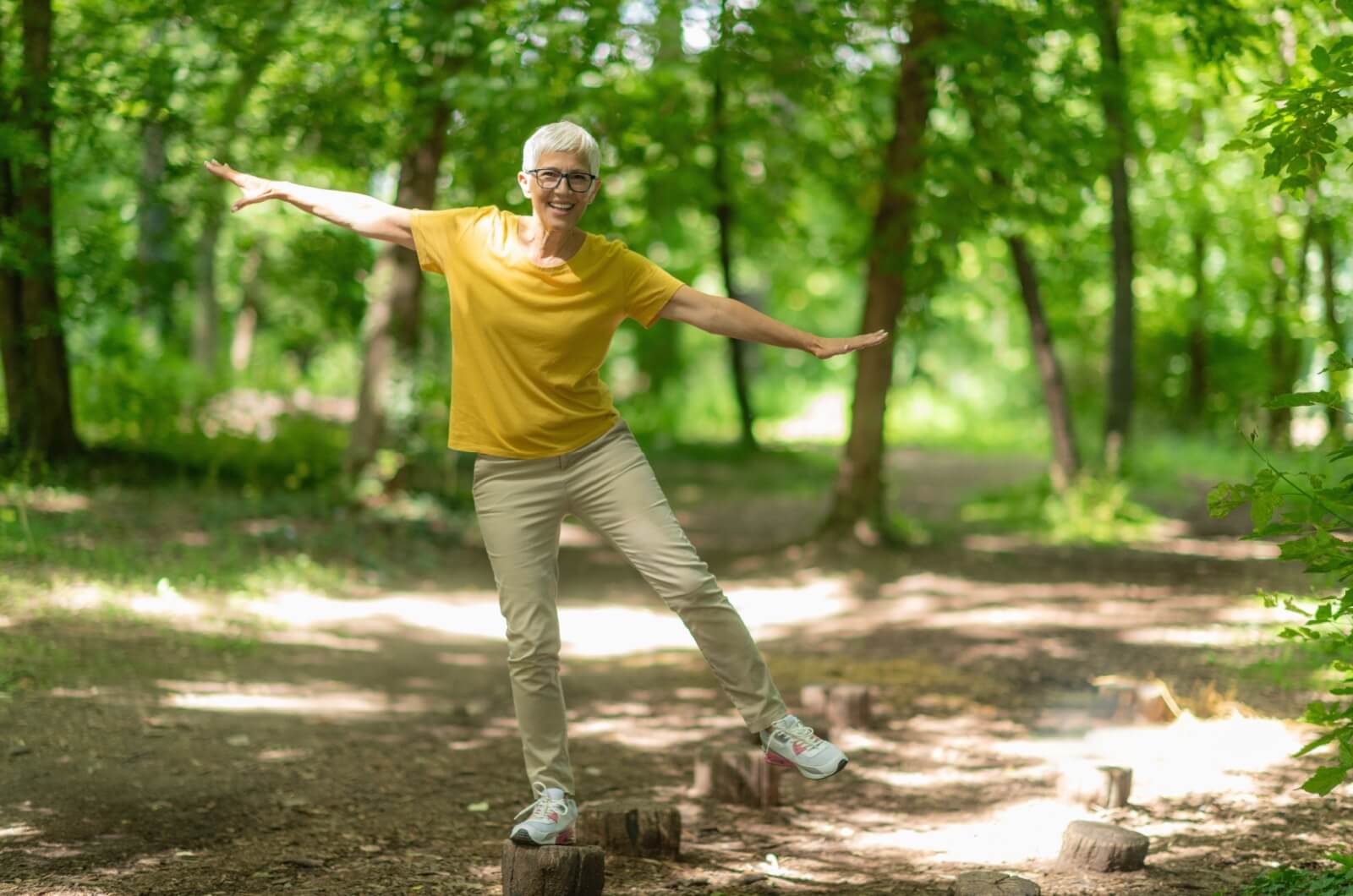 A smiling senior outdoors with trees in the background balancing with their right leg on a tree stump and arms to their sides.