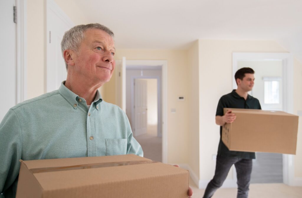 Older man smiling while carrying a moving box, assisted by another person in a new room.