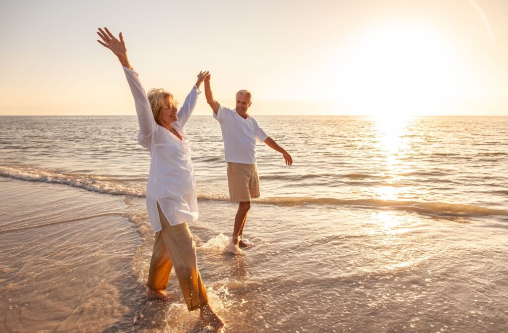 Two happy seniors walking on a beach, holding hands with one hand up in the air.