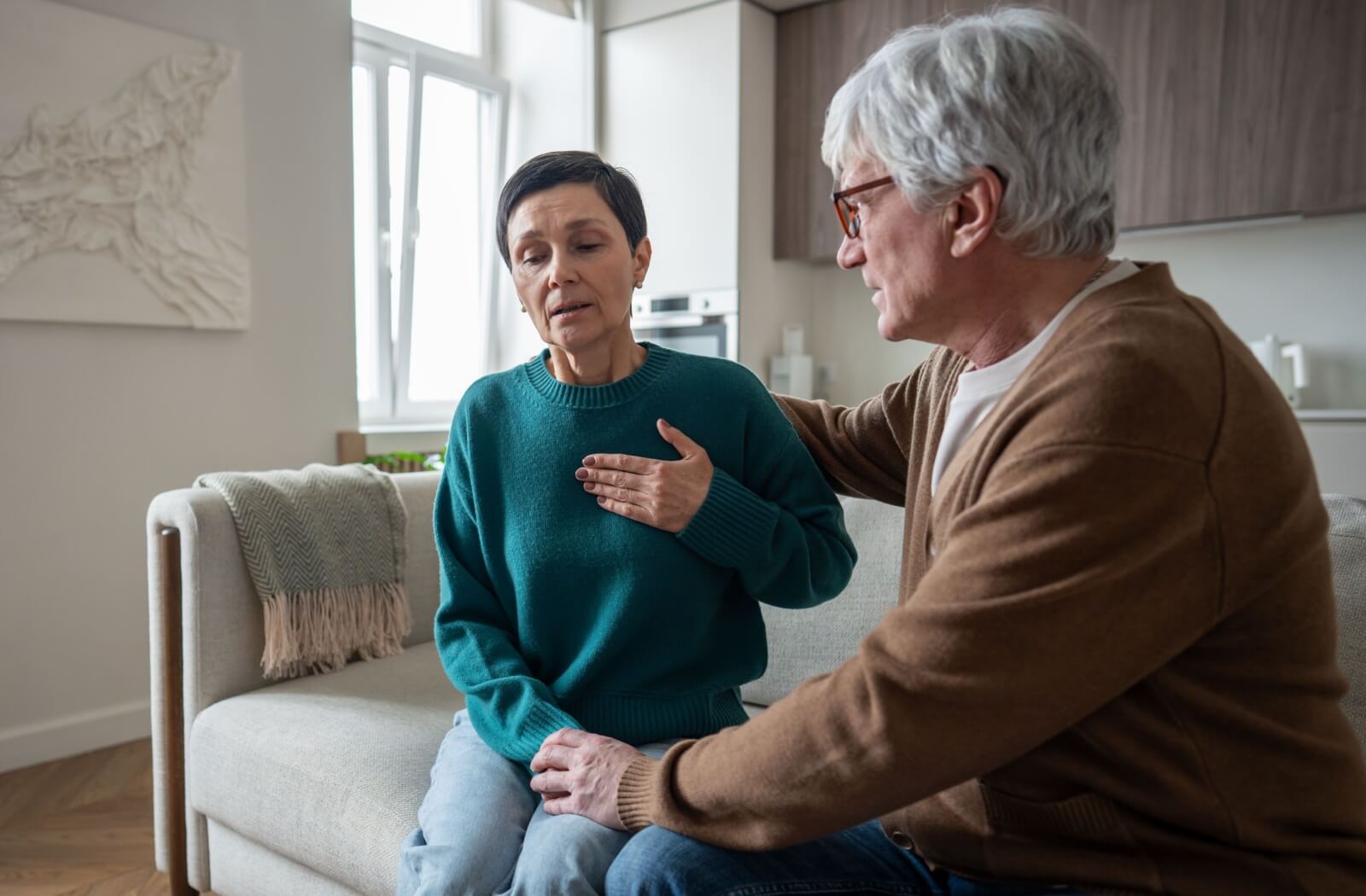 Older adult sitting on a couch with hand on chest while another person offers comfort with a hand on their shoulder