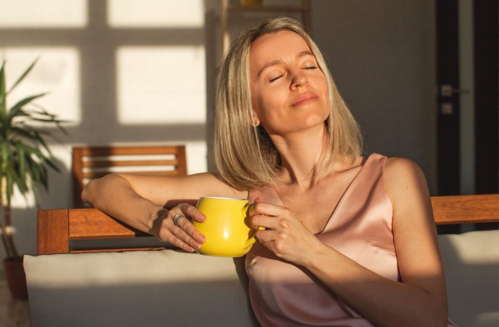A middle-aged adult sitting in their sunlit living room and drinking tea to practice mindfulness and prevent burnout.