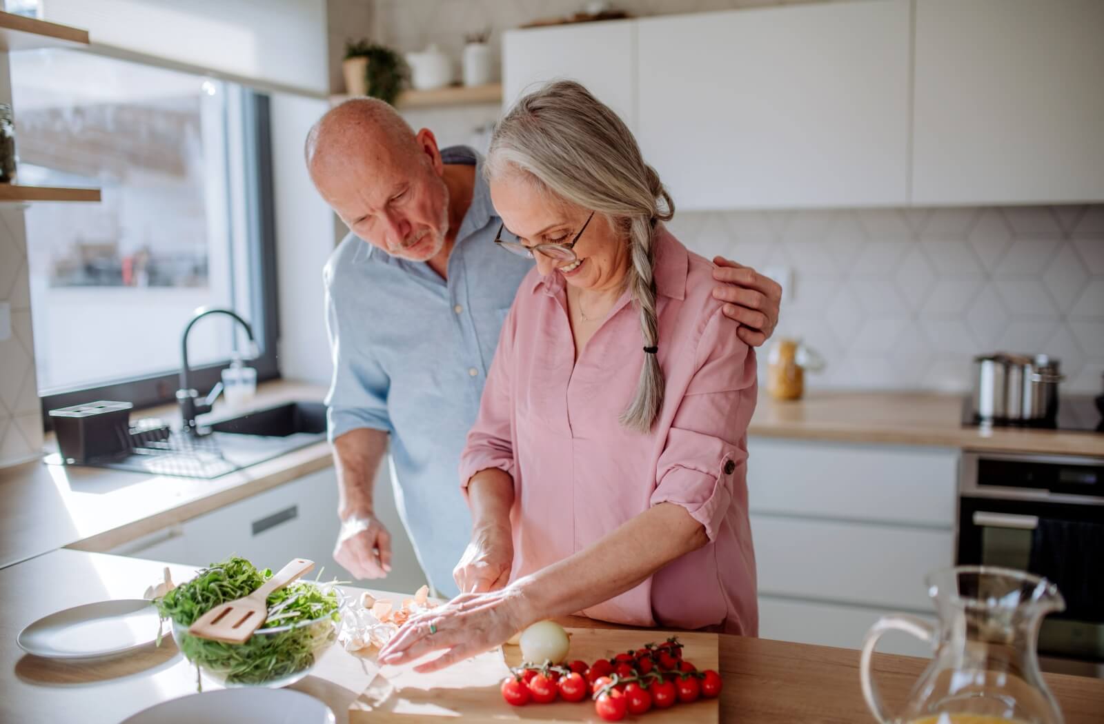 Older couple cooking together in a bright kitchen with fresh vegetables and greens on the counter