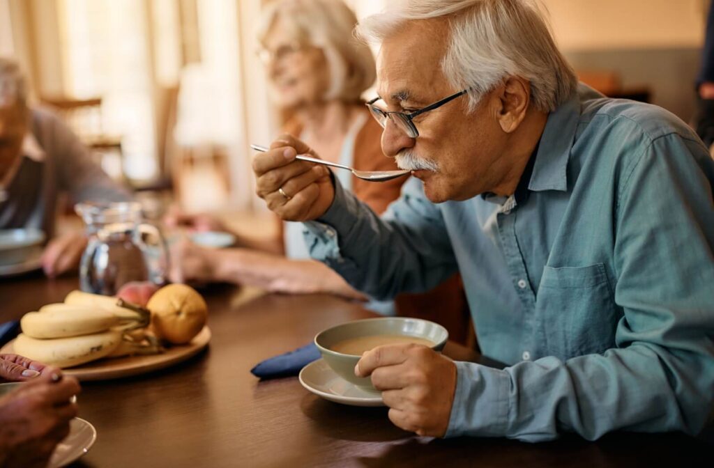 A senior takes a sip from a bowl of soup offered during a mealtime in senior living, while surrounded by friends