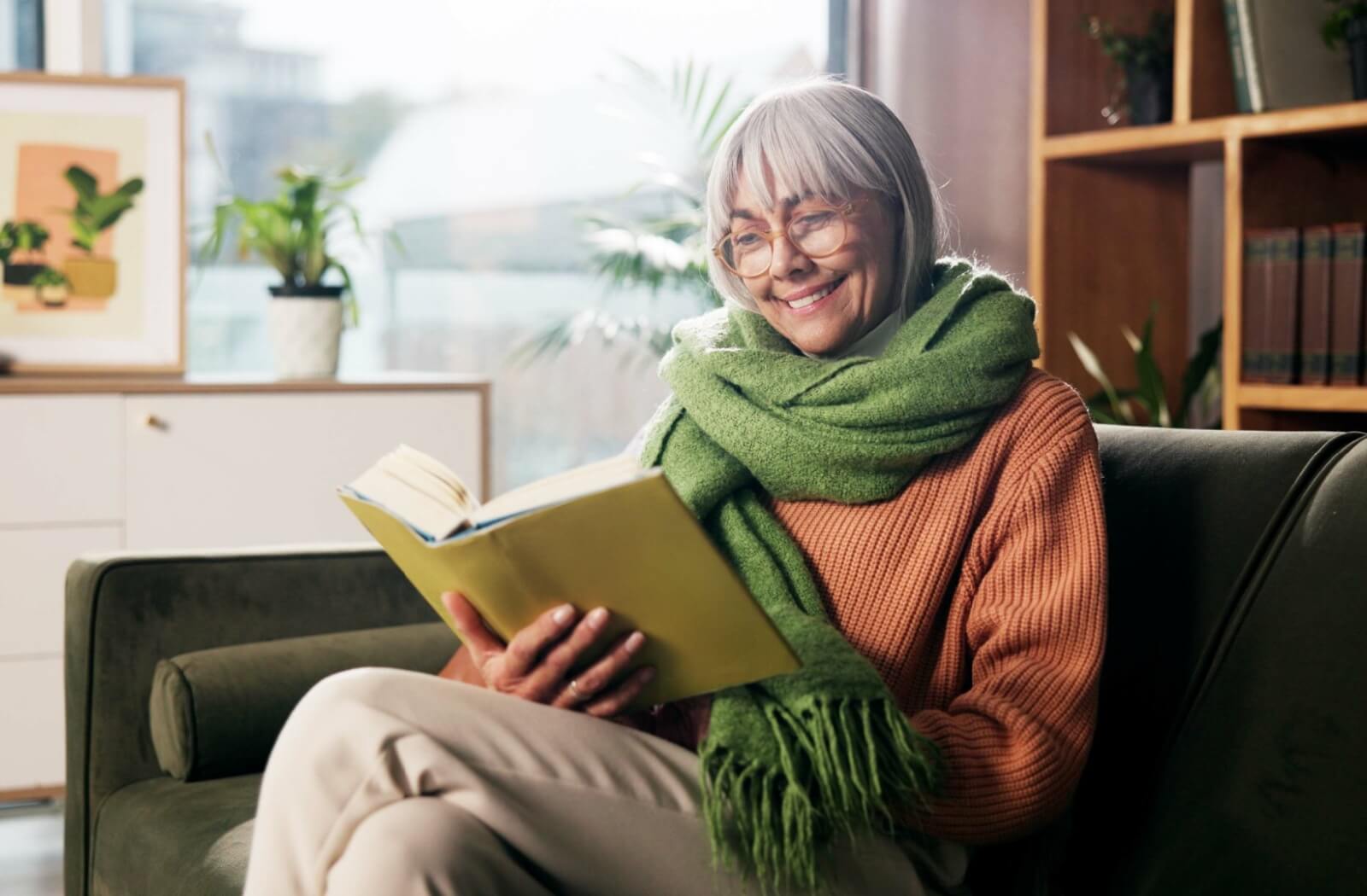 Older adult with gray hair and glasses sitting on a sofa reading a book with a green scarf wrapped around their shoulders