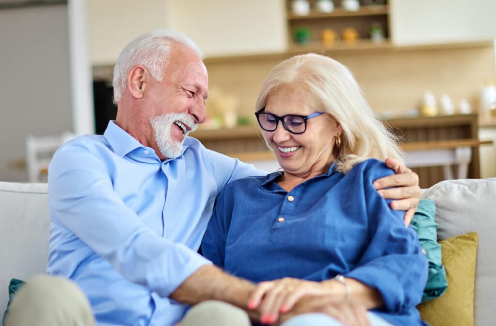 Senior couple sitting on a couch smiling and laughing together in a cozy assisted living apartment